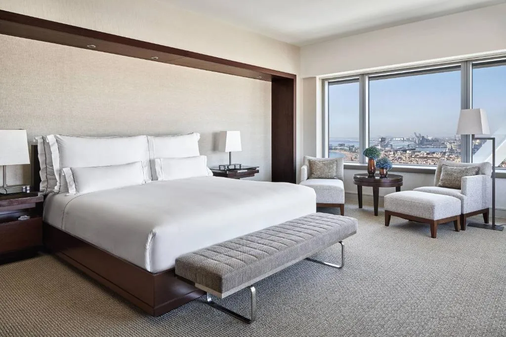 habitación con cama para dos, sillas cómodas y un amplio ventanal que ofrece vistas impresionantes tanto de la ciudad como del mar.