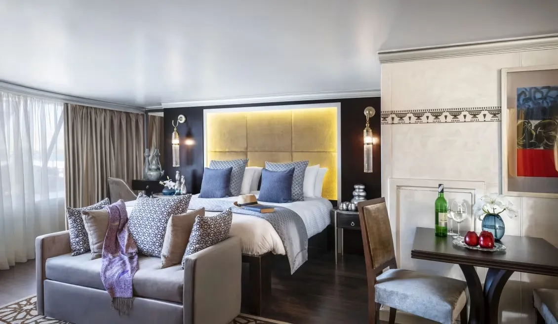 En la habitación para dos personas, encontrarás una cama amplia, un sofá cómodo, un comedor con mesa para dos personas. Sobre la mesa, se ofrecen tres manzanas frescas, dos copas de vino y una botella de vino