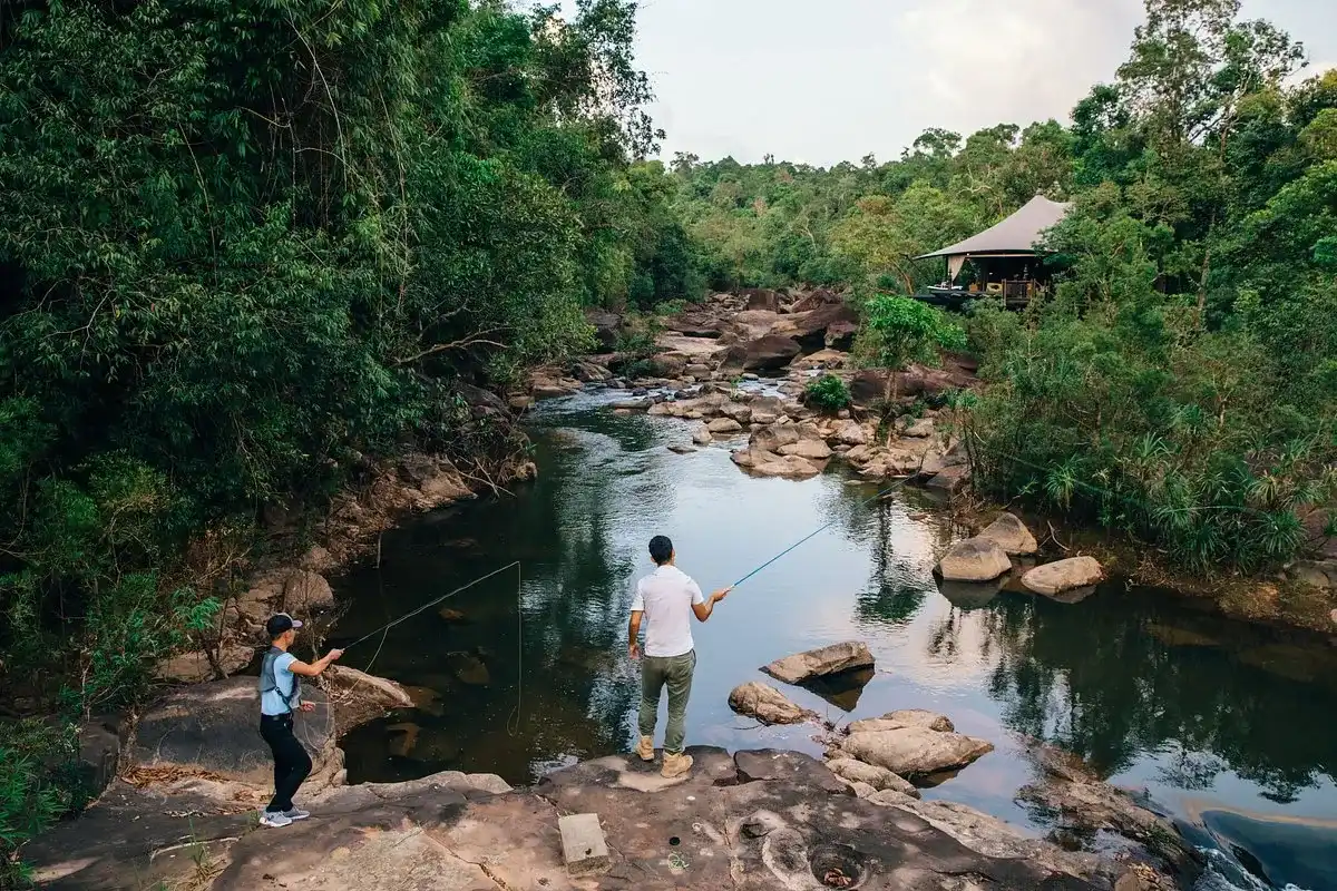 Dos turistas disfrutando de una experiencia única mientras pescan en el río, rodeados de árboles y de zona verde en Shinta Mani salvaje.