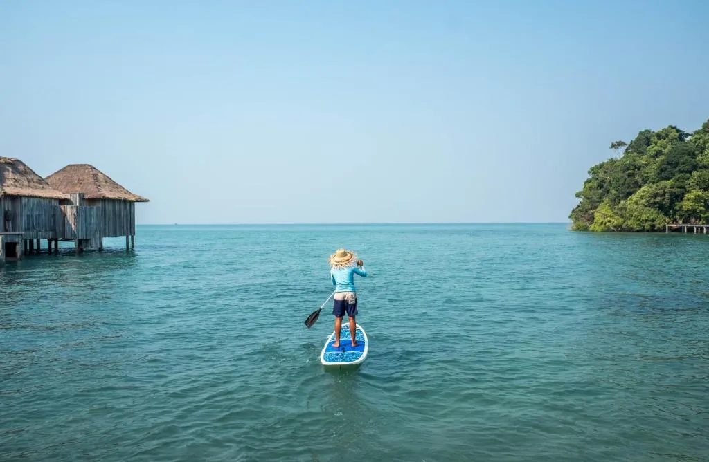 turista disfrutando de una experiencia única en Song Saa Island, montado en una tabla de surf y remando suavemente en el mar cristalino. La isla ofrece un entorno perfecto para los amantes de los deportes acuáticos