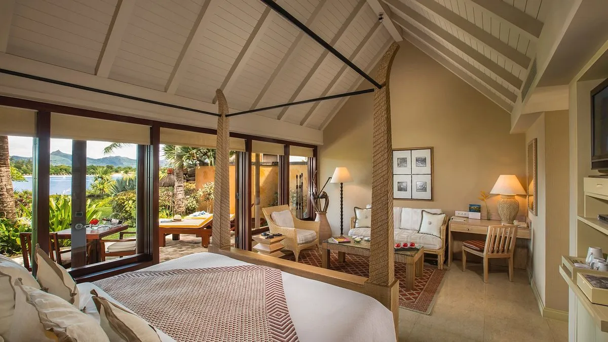 habitación grande cuenta con una cama para dos personas, televisor, escritorio con lampara y una silla, sillones, una mesa decorativa, ventanal que conecta con el el jardín privado con vista al mar