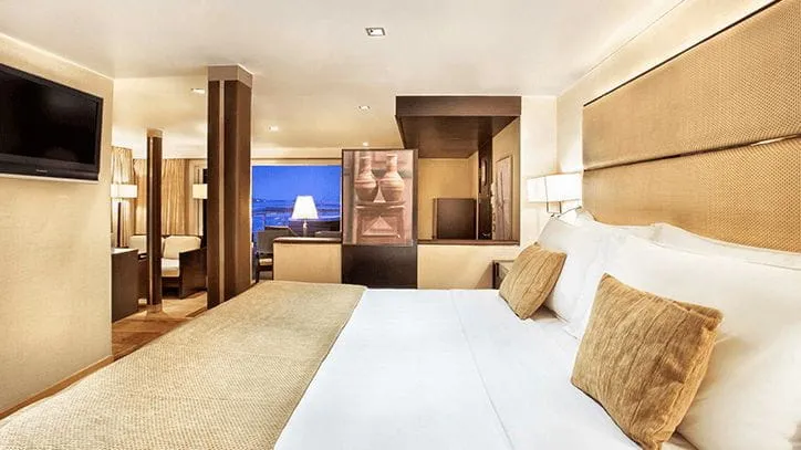 habitación de lujo, con una cama para dos personas, televisor, asiento, al lado, en la misma habitación, hay un sillón espacioso con una mesa decorativa y un ventanal con vista al mar.