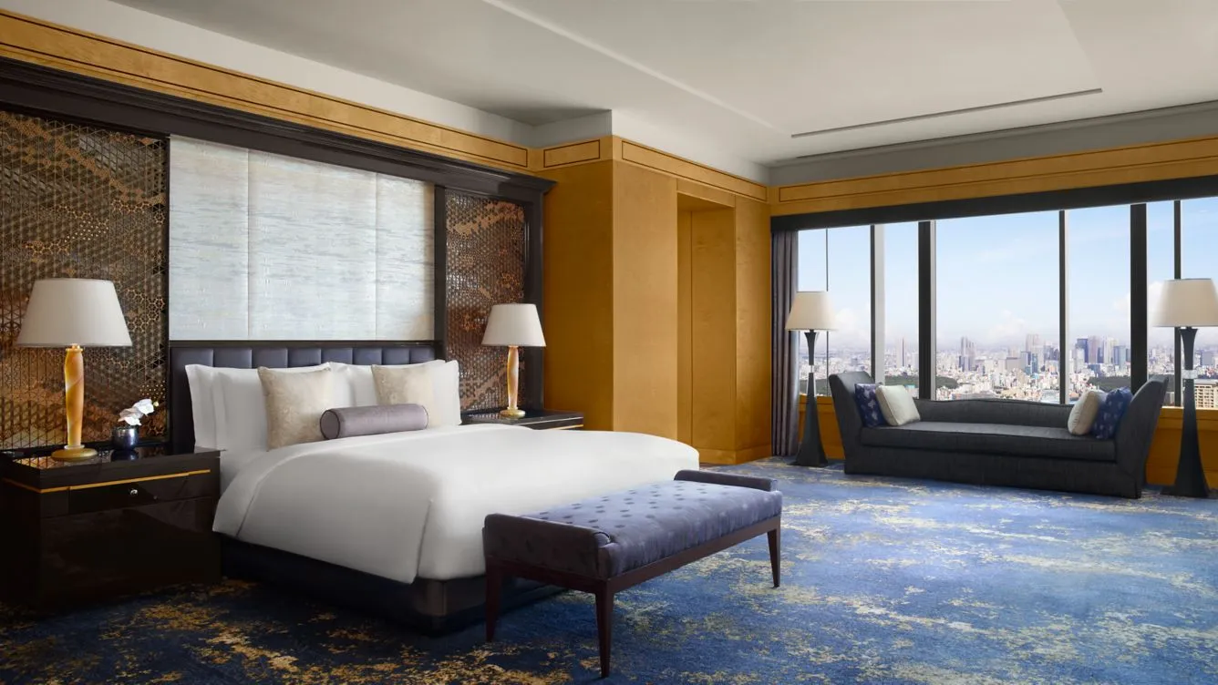 habitación de lujo con cama para dos personas, nocheros cada uno con una lampara, sillón para una mejor comodidad y una ventana con vista a la ciudad.