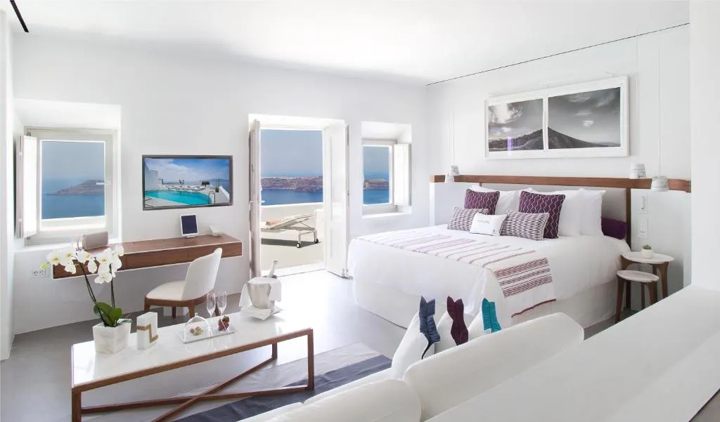 suite de color blanca con cama amplia, escritorio de color café y sillón cómodo, con puerta al frente para que puedas salir al balcón y ver la increíble vista al mar en Santorini.