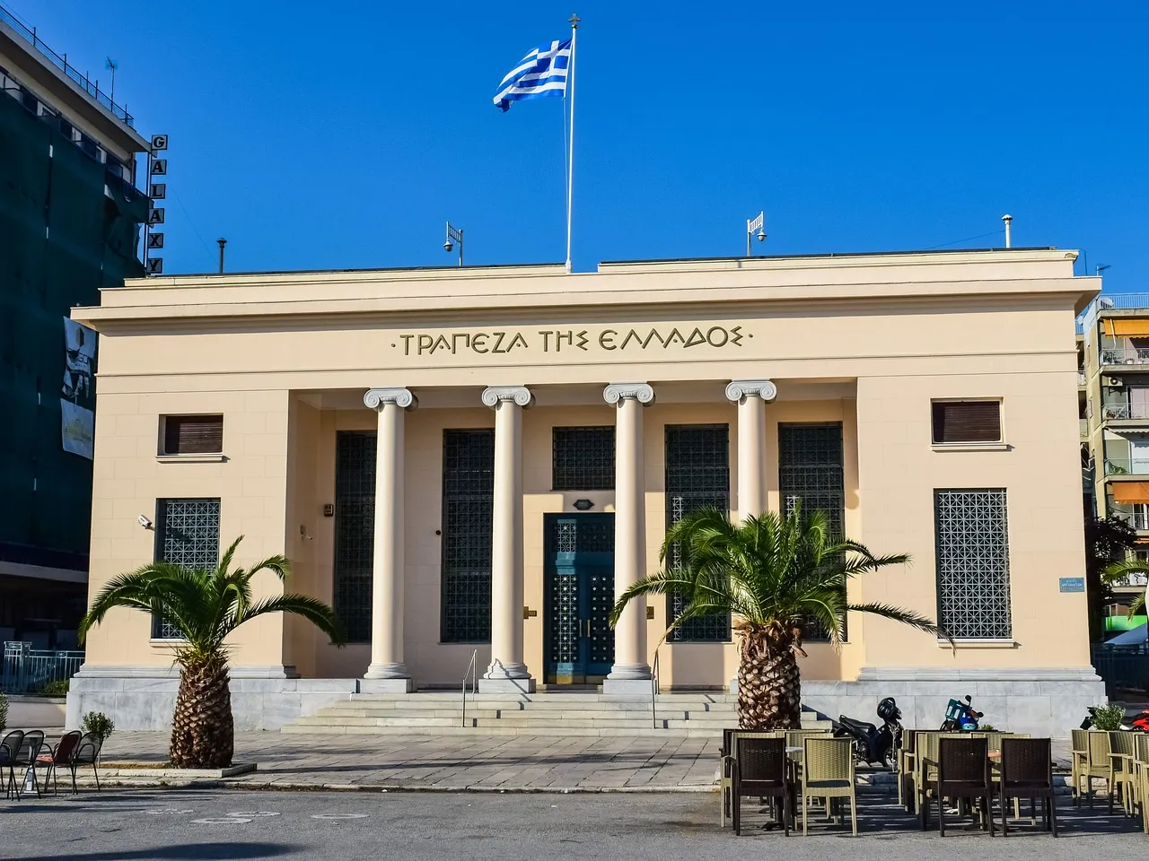 edificio de color beige con la bandera de Grecia, rodeado de otros edificios, palmeras y sillas un lugar emblemático para conocer.