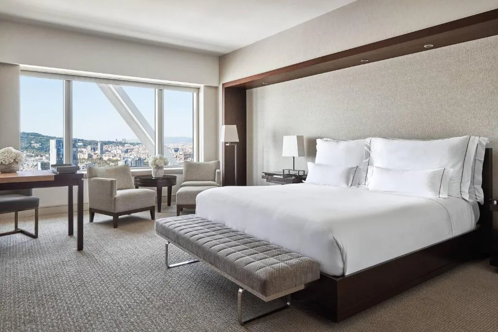 habitación con cama para dos personas, escritorio, sillas cómodas y un amplio ventanal con vista a la ciudad.